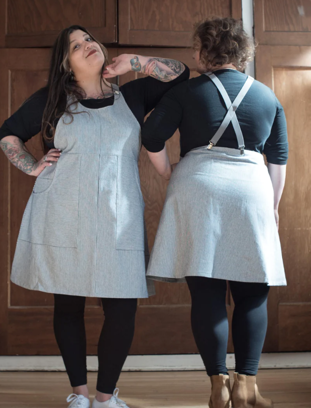 Image description: Two fat people, one facing the camera, one facing away, wear the same pinafore style dress in a black and white railroad stripe pattern