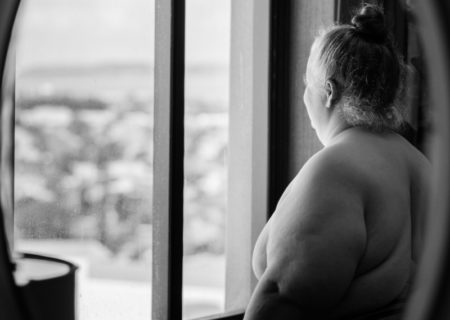 In this black and white photo, a fat white woman stands nude in front of a window high above a city and looks out, reflected in an oval mirror.