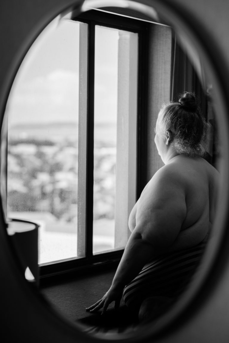 In this black and white photo, a fat white woman stands nude in front of a window high above a city and looks out, reflected in an oval mirror.