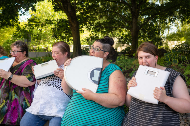 Four fat people in summer clothing hold bathroom scales and playfully bite them.