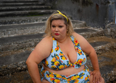 A fat white woman with blonde hair sits on concrete water stairs in a blue and fruit-print bikini.