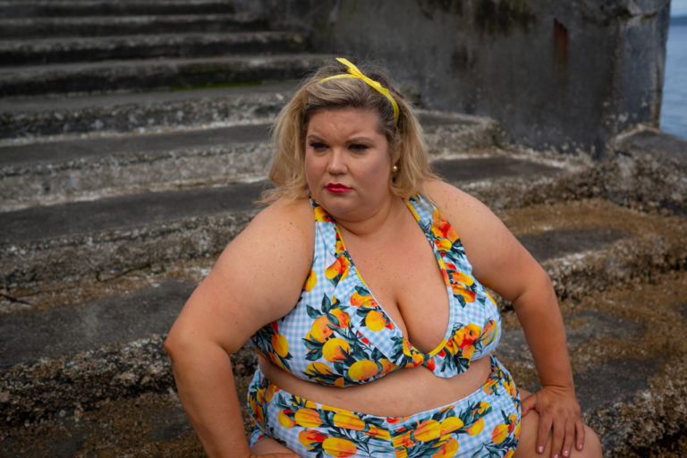 A fat white woman with blonde hair sits on concrete water stairs in a blue and fruit-print bikini.