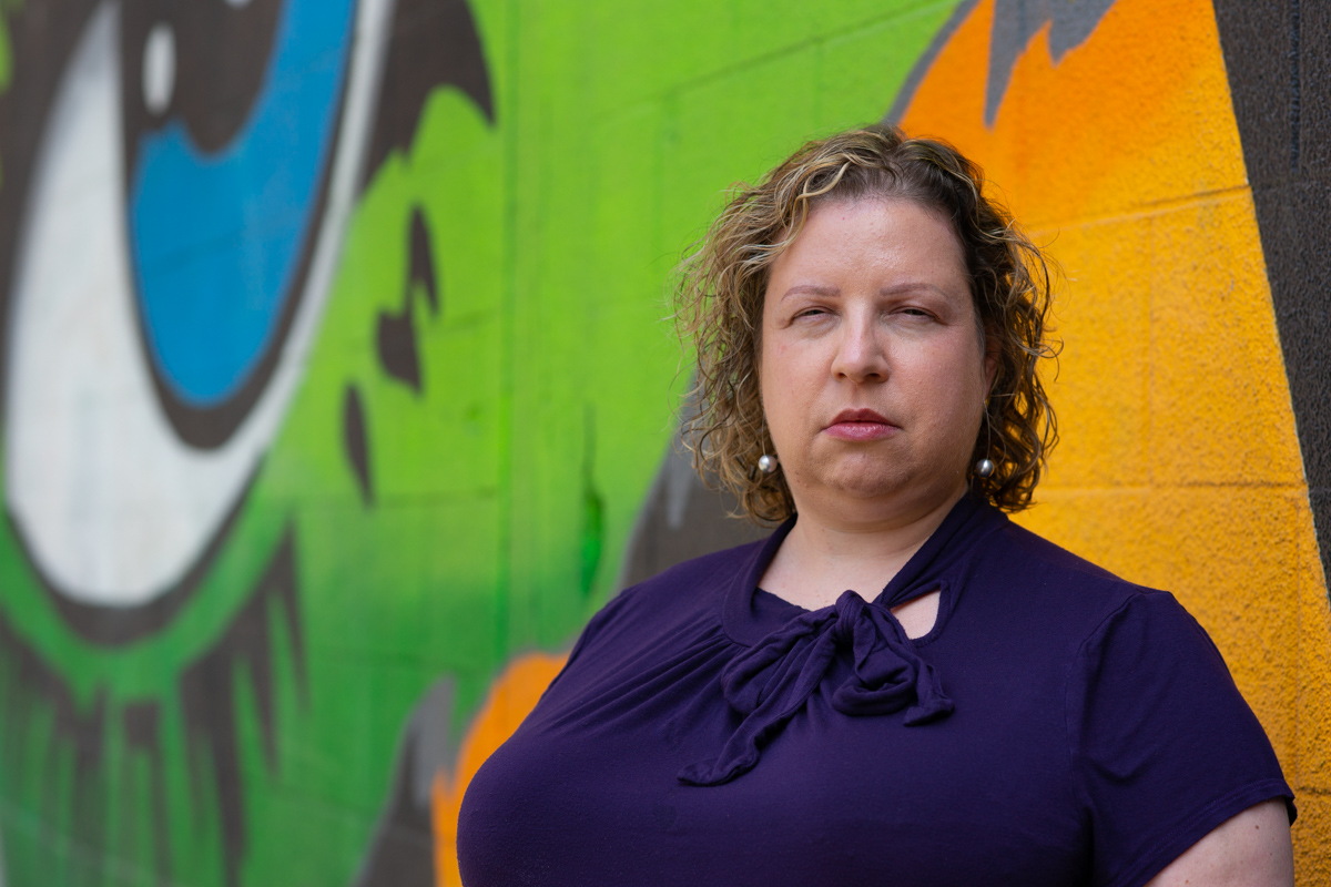A woman with short curly hair, wearing a purple short-sleeved top, stands in front of a colorful mural with a neutral expression. She's gearing up to ask someone to stop dieting or being fatphobic.