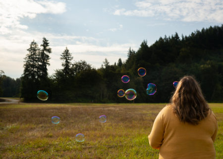 A fat woman with long brown hair and a yellow sweater blowing bubbles in a field. Her back is to the camera.