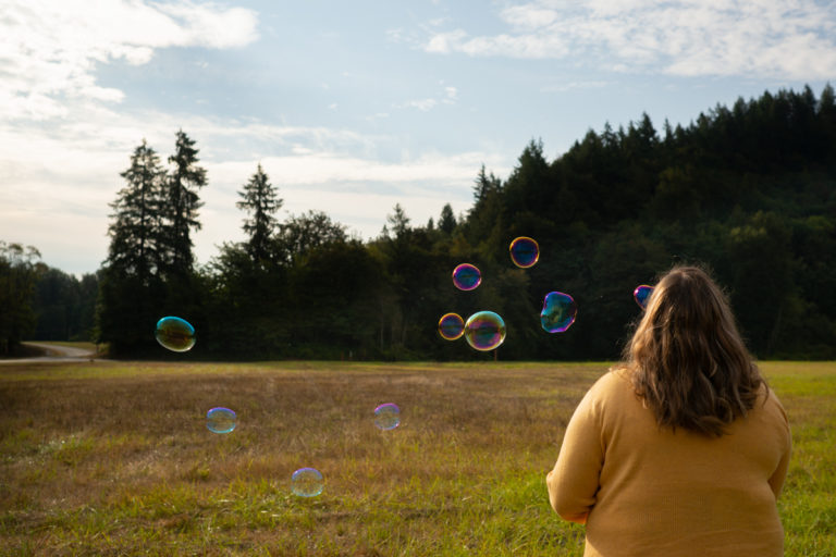 A fat woman with long brown hair and a yellow sweater blowing bubbles in a field. Her back is to the camera.