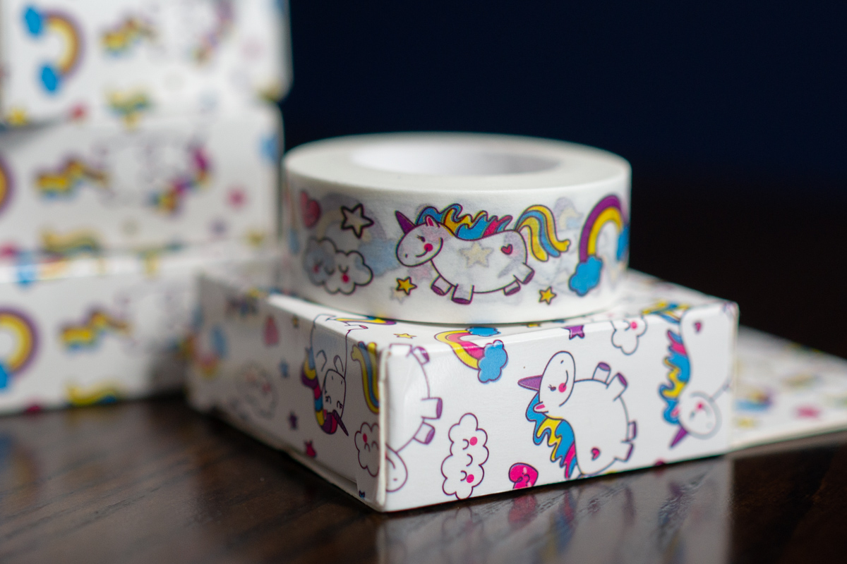 A roll of white washi tape with a cute unicorn design sitting on a similarly-patterned box, with a stack of more boxes in the background.