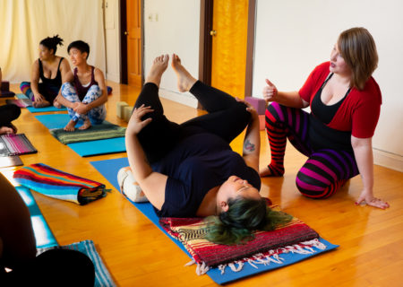 A set of photos depicting a yoga class full of students in a studio with red and white walls and a wooden floor. The students are in various yoga poses, meditating and being assisted by an instructor