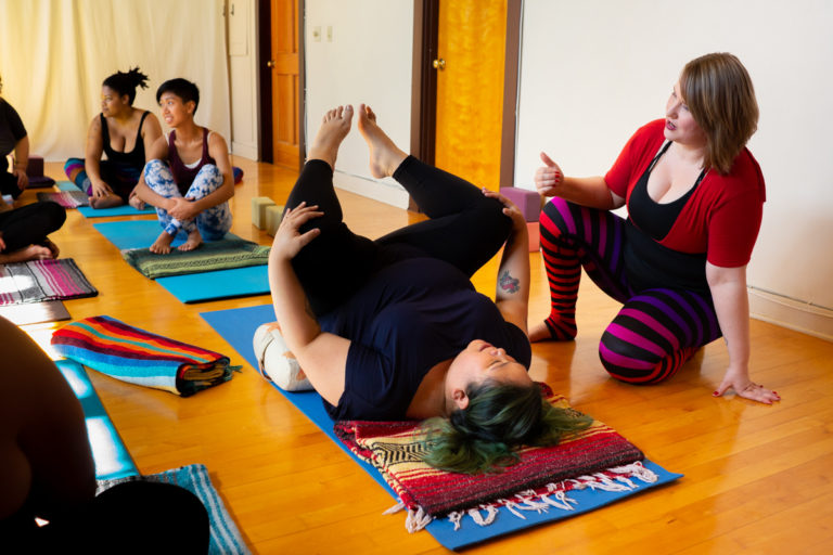 A set of photos depicting a yoga class full of students in a studio with red and white walls and a wooden floor. The students are in various yoga poses, meditating and being assisted by an instructor