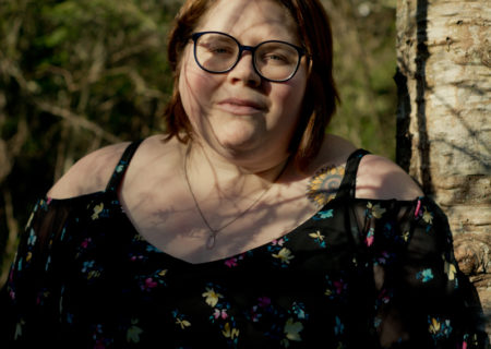 A fat white woman with glasses, short brown hair and a floral cold-shoulder top stands in a forest with shadows from tree branches crossing her face and a neutral expression.