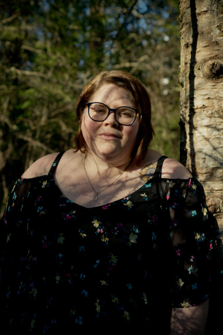 A fat white woman with glasses, short brown hair and a floral cold-shoulder top stands in a forest with shadows from tree branches crossing her face and a neutral expression.