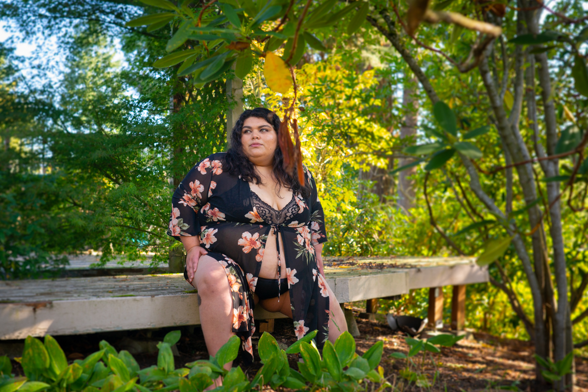 A fat woman with brown skin and black hair sits in a bra, panties and flowing robe on the edge of a wooden deck, surrounded by green plants, at a Seattle body-positive botanical boudoir photo session.