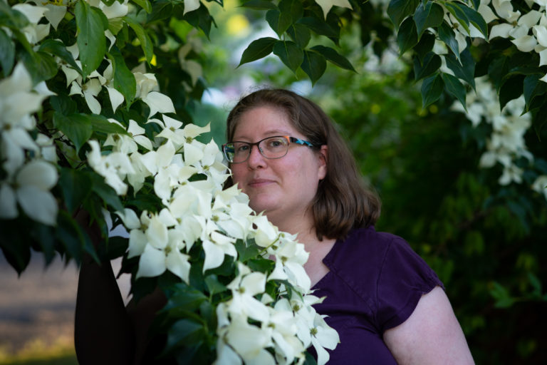 Lindley, a fat white woman with shoulder-length brown hair and glasses, stands in a garden at dusk holding a branch laden with white dogwood flowers.