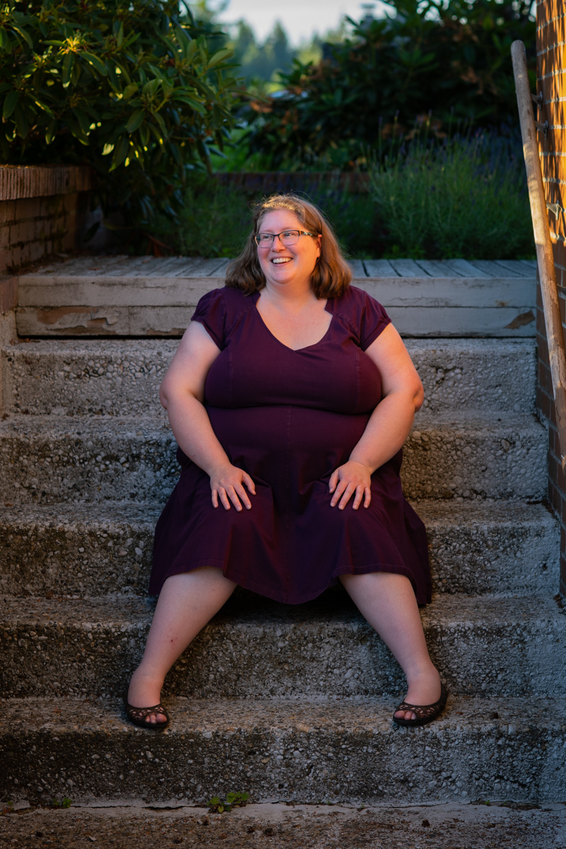 A fat white woman celebrating her 20th wedding anniversary sits on a set of gray steps, wearing glasses and a purple dress.