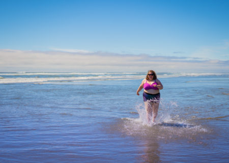 Lindley, a fat white woman in sunglasses and a two-piece swimsuit, splashes through shallow ocean water on a sunny day.
