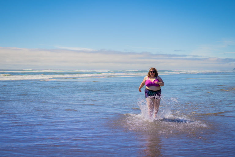 Lindley, a fat white woman in sunglasses and a two-piece swimsuit, splashes through shallow ocean water on a sunny day.