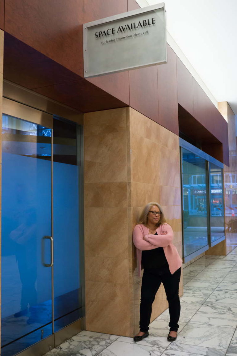 A Black female entrepreneur stands in an office building under a sign for a retail space for rent.