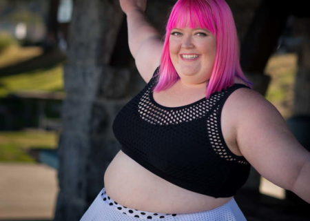 A fat woman with bright pink hair, wearing a black crop top and white skirt, smiles at the camera with her arms outstretched outdoors in a park.