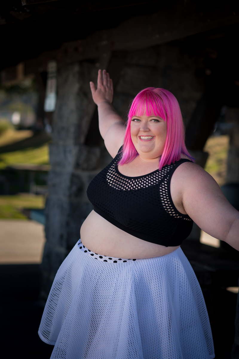 A fat woman with bright pink hair, wearing a black crop top and white skirt, smiles at the camera with her arms outstretched outdoors in a park.