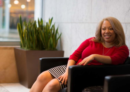 A plus-size Black woman in a striped skirt and red sweater sits in a leather chair in an office lobby, looking off to one side and smiling slightly.