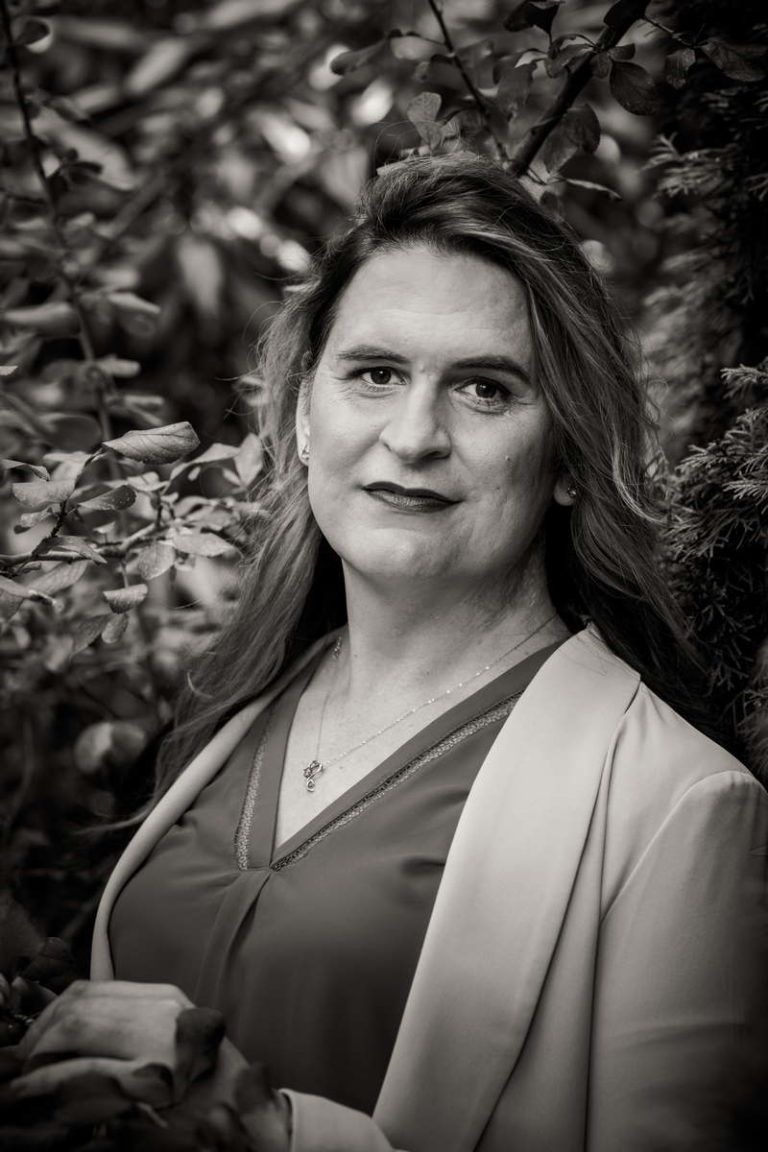 A black-and-white photo of a trans woman with long hair. She is wearing a v-neck blouse, blazer and necklace and is smiling gently in front of bushes.