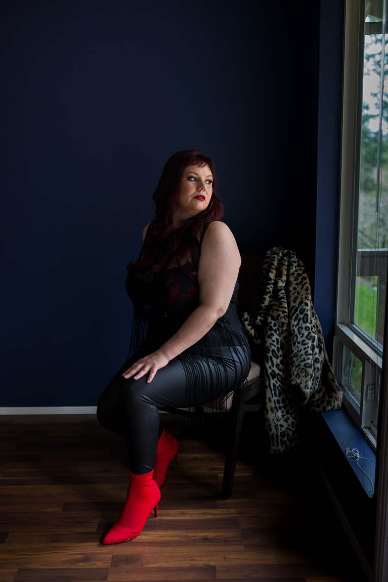 A woman with pale skin, black clothing and bright red boots sits in a chair in a darkish room, looking over her shoulder out the window. A leopardskin coat hangs from the chair back. She's at a body positive, plus size boudoir session in Seattle.