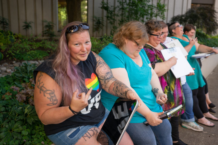 Five fat people in casual clothing sit on a concrete wall in front of bushes and a building. They're all holding bathroom scales and giving the scales a middle finger.