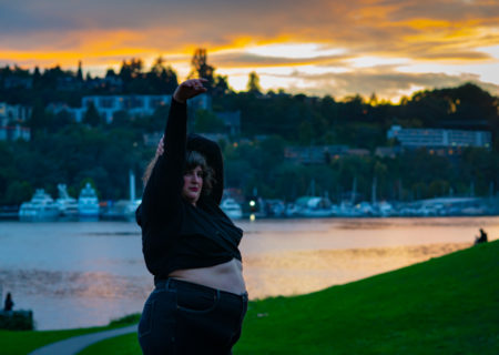 A fat white woman in a cropped sweater and jeans stands with one arm in the air, one wrapped around her head and her belly exposed in a park with green grass at sunset with water and houses behind her.