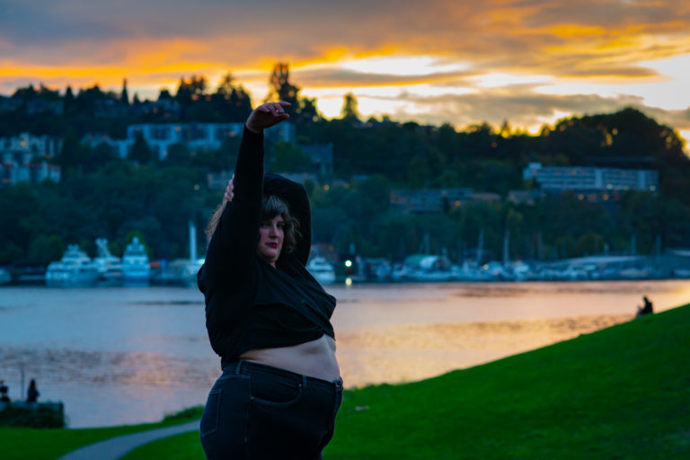 A fat white woman in a cropped sweater and jeans stands with one arm in the air, one wrapped around her head and her belly exposed in a park with green grass at sunset with water and houses behind her.