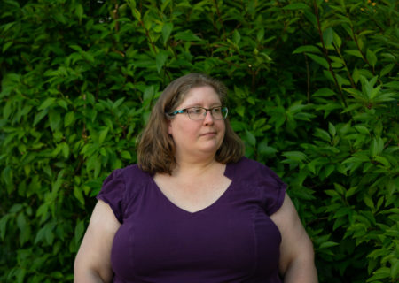 Lindley, a fat white woman in a purple dress and glasses, stands in front of green bushes and looks away from the camera. She looks annoyed, resigned or irritated.