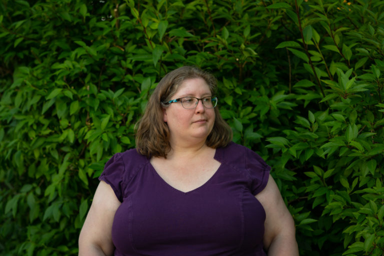 Lindley, a fat white woman in a purple dress and glasses, stands in front of green bushes and looks away from the camera. She looks annoyed, resigned or irritated.