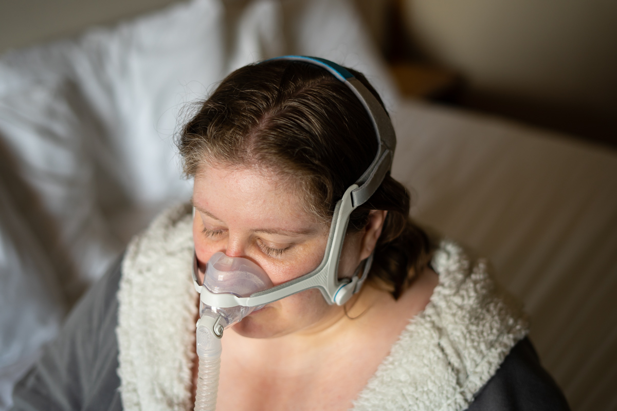 Lindley, a fat white woman in a fluffy gray and white robe, is sitting on a bed with her CPAP headgear and mask on and her eyes closed.