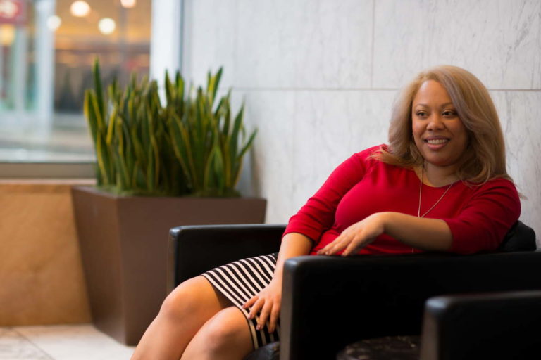 A plus-size Black woman in a striped skirt and red sweater sits in a leather chair in an office lobby, looking off to one side and smiling slightly.