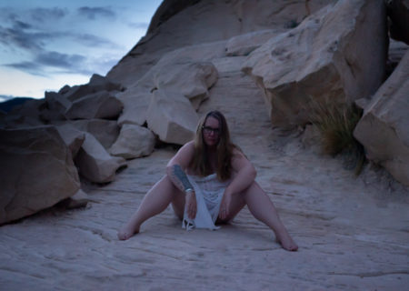 A young woman draped in a white scarf sits on a rocky ridge at twilight during a body positive photo shoot.