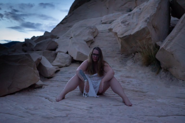 A young woman draped in a white scarf sits on a rocky ridge at twilight during a body positive photo shoot.