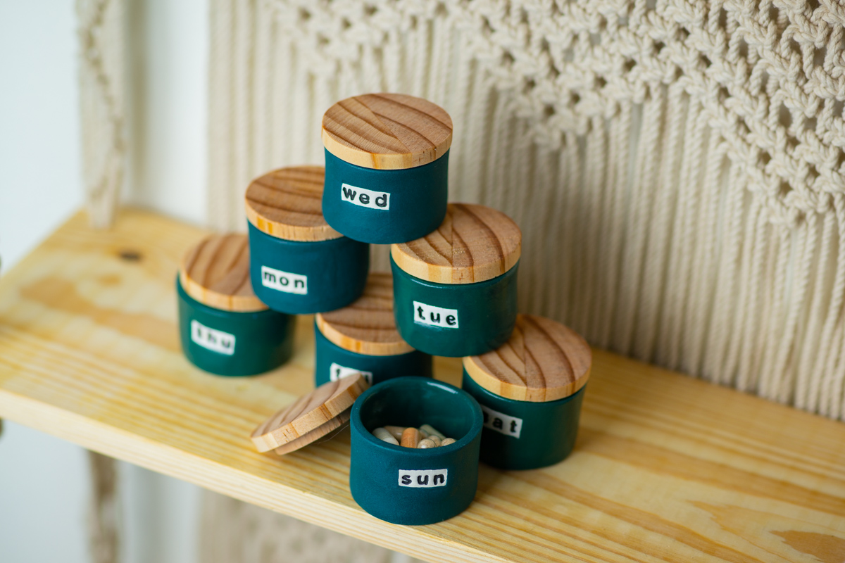 A set of small, dark teal pottery vessels with wooden lids sits on a wooden shelf held up by cream-color macrame weaving. One of the vessels contains a variety of pills.