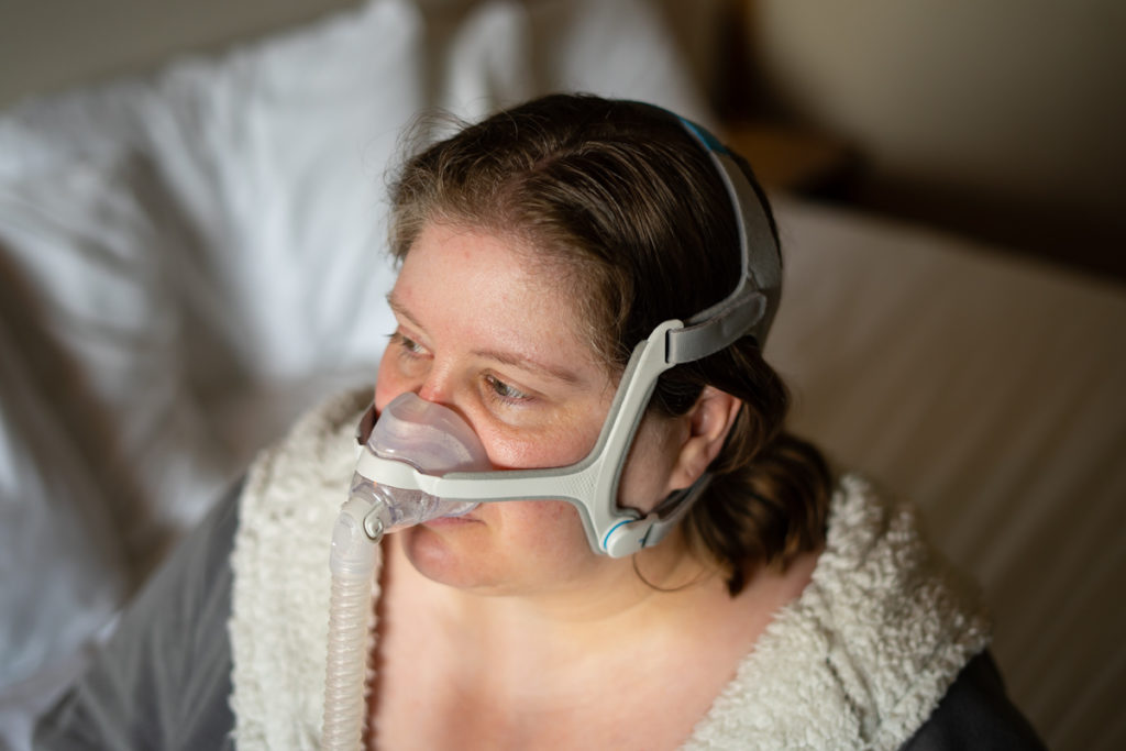 A plus-size white woman wears a fuzzy robe and a CPAP device and looks to one side in a weight-neutral healthcare stock photo.
