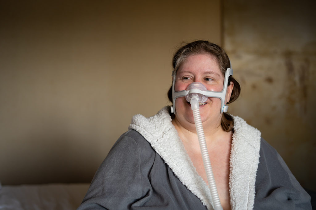 A plus-size white woman wears a fuzzy robe and a CPAP device and looks to one side in a weight-neutral healthcare stock photo.