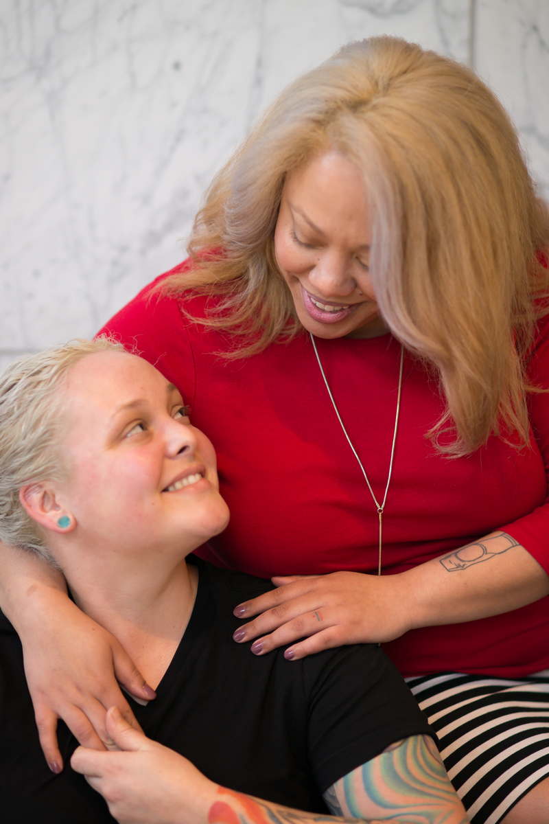 A close-up photo of a married couple smiling at each other. One is a Black woman in a red top and striped skirt. The other is a white woman in a black shirt with colorful tattoos.