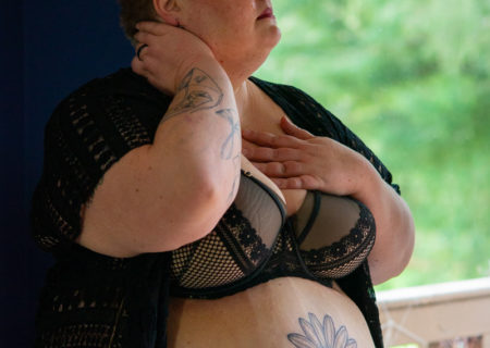 a fat woman with very short hair and arm and belly tattoos has her hands on her neck and chest and is standing just inside a large window looking out. She's wearing a black bra, pink panties and black robe and has a neutral expression.