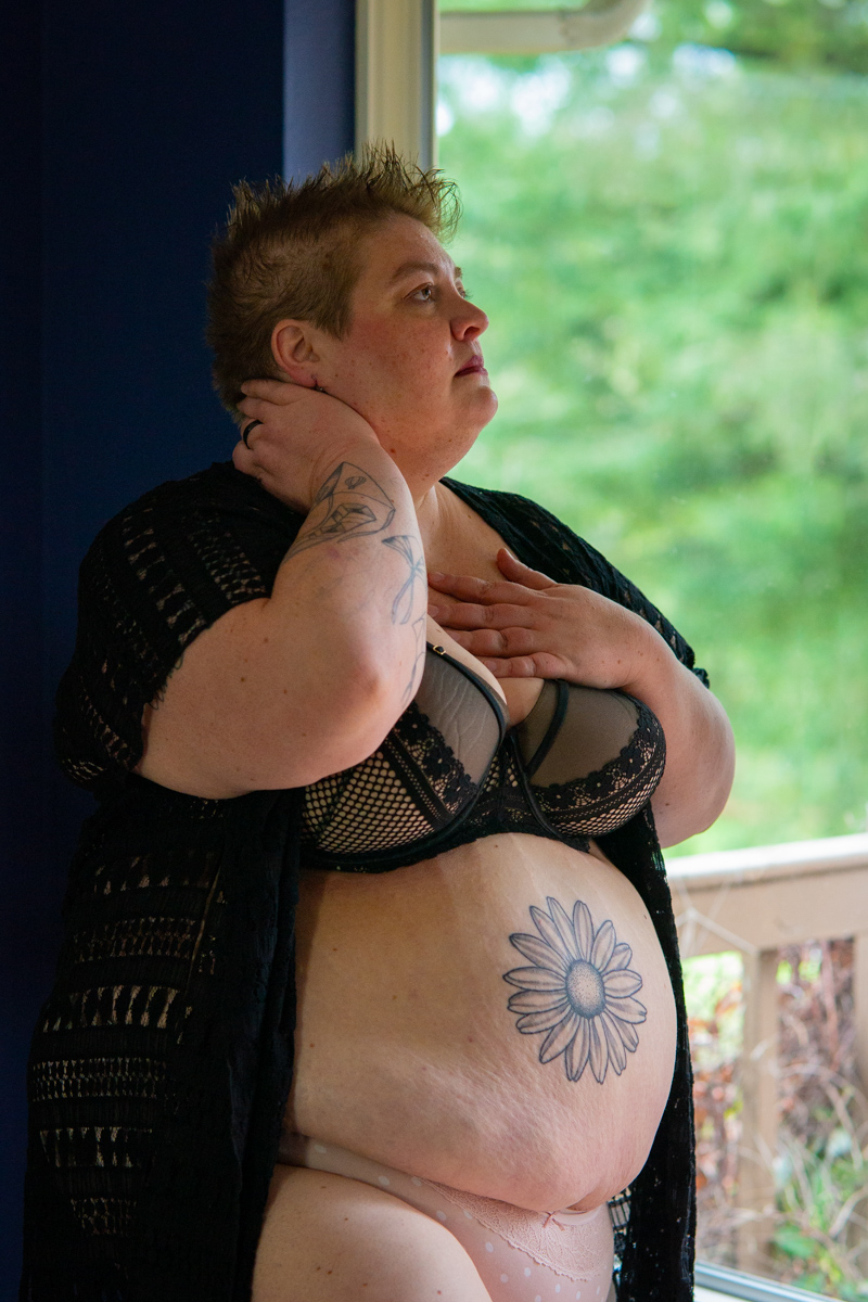 a fat woman with very short hair and arm and belly tattoos has her hands on her neck and chest and is standing just inside a large window looking out. She's wearing a black bra, pink panties and black robe and has a neutral expression.