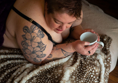 A fat white woman with short hair lies under blankets at a body positive boudoir shoot in Seattle, WA. She's holding a coffee cup, looking down and smiling.