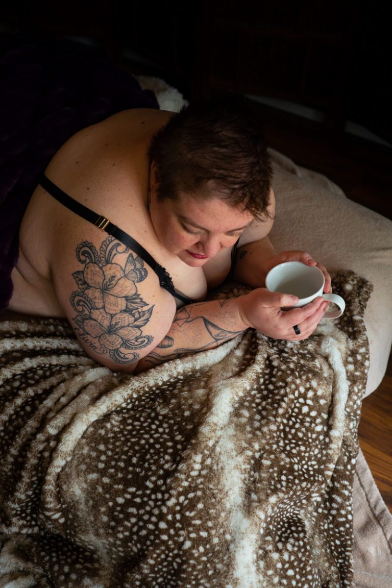 A fat white woman with short hair lies under blankets at a body positive boudoir shoot in Seattle, WA. She's holding a coffee cup, looking down and smiling.