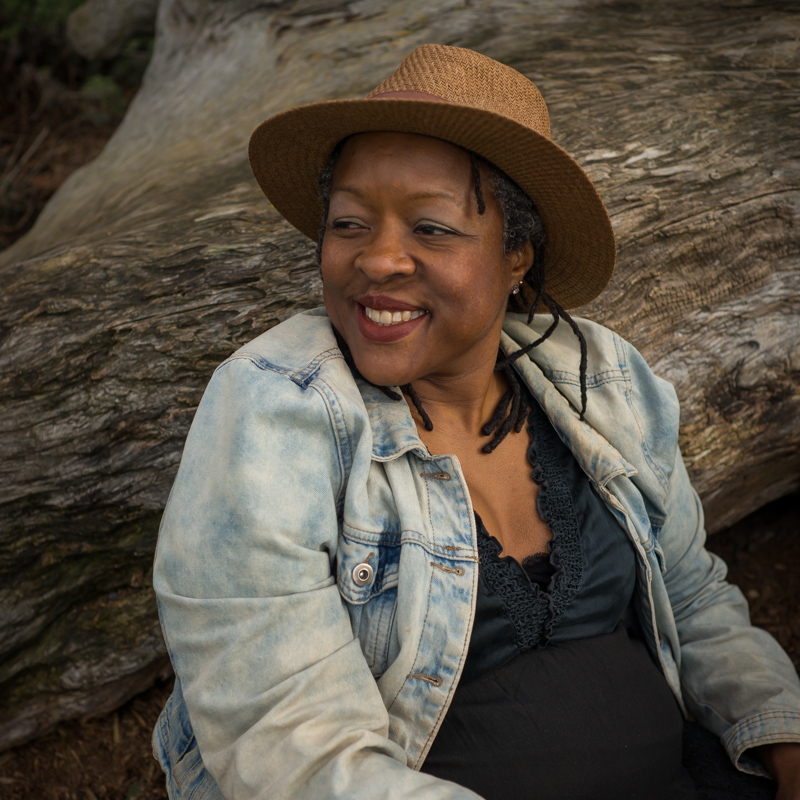 A Health at Every Size stock photo featuring a Black woman sitting outdoors in a hat and jacket, leaning against a log.