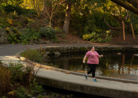 A fat white woman in a pink shirt and black pants runs along a paved pathway around a pond in a park.