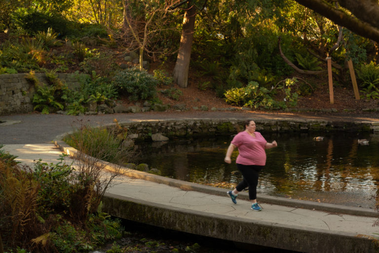 A fat white woman in a pink shirt and black pants runs along a paved pathway around a pond in a park.