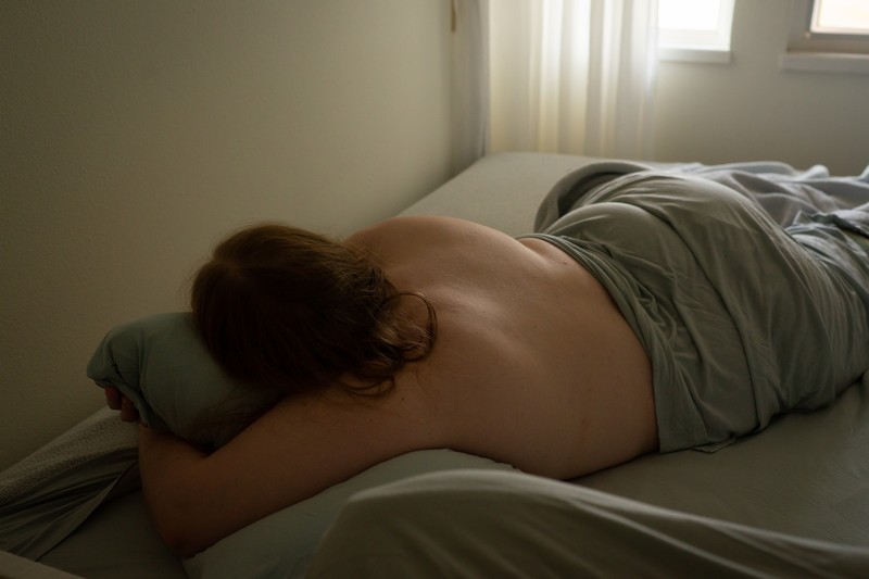 A plus-size woman lies in a bed in a white room with pale blue sheets, a window and white curtains. She may be sleeping, napping, depressed, or just resting.