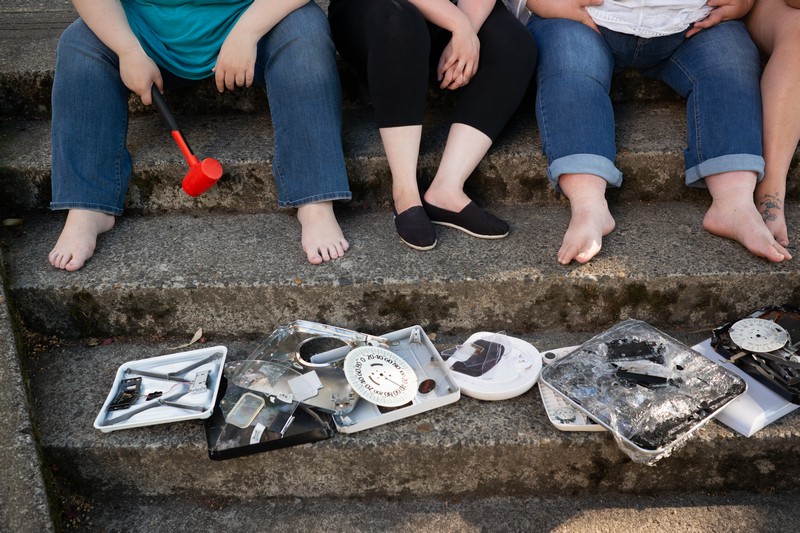 A pile of smashed and broken bathroom scales lies on a concrete step in an urban park. Several women's feet are resting on the next step up.