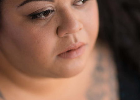 A fat woman of Hawaiian and Tongan heritage is shown from the bust up, looking away from the camera with a pensive expression. She's wearing a black v-neck top and has traditional-style tattoos on her chest.