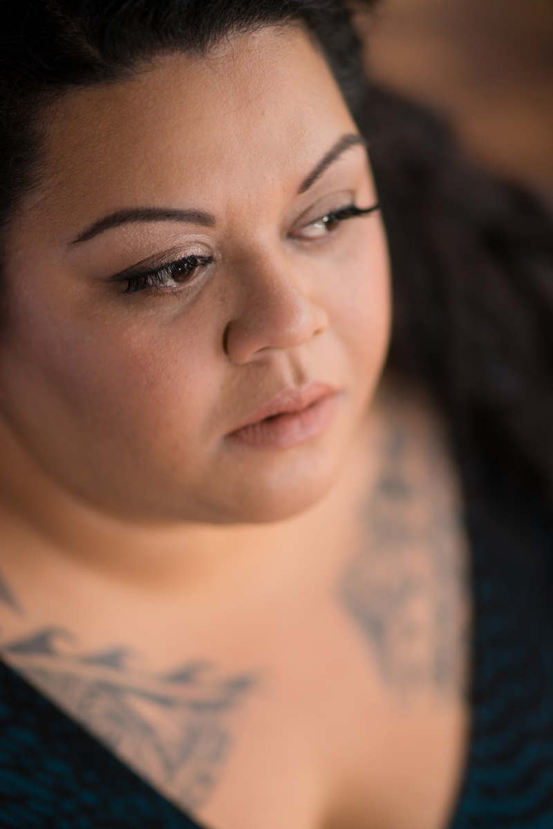 A fat woman of Hawaiian and Tongan heritage is shown from the bust up, looking away from the camera with a pensive expression. She's wearing a black v-neck top and has traditional-style tattoos on her chest.