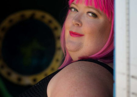 A close-up photo of a fat white person with bright pink hair and lipstick, looking over their shoulder towards the camera. They're wearing a sleeveless black top and leaning against a wall with a nautical-style porthole in the background.